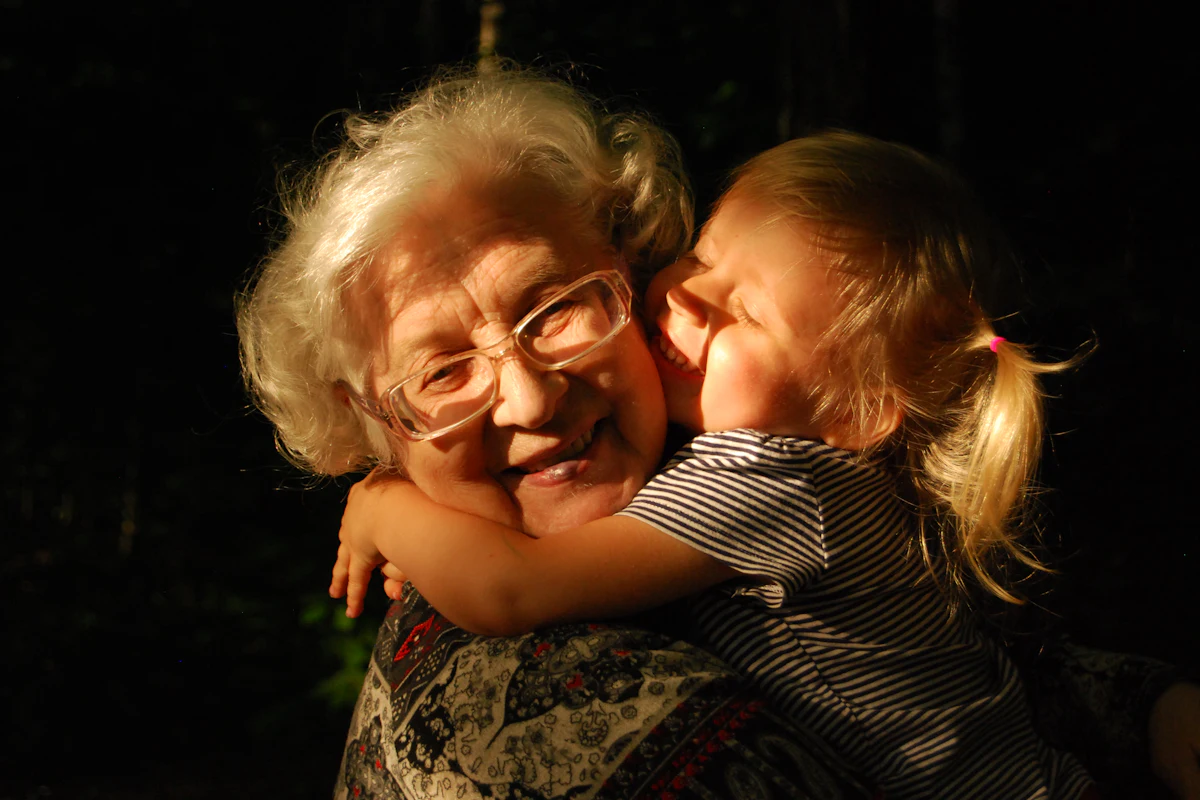 A grandmother with white hair and glasses hugs her grandchild tight, both smiling.