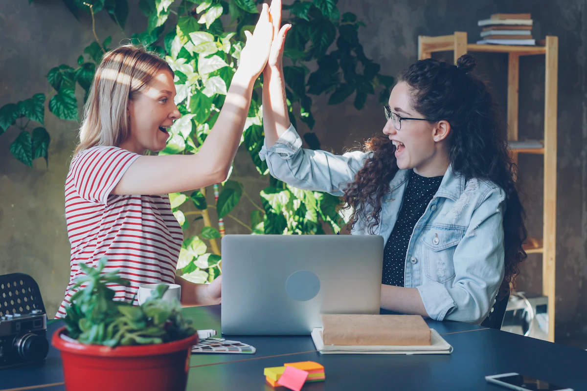 Two colleagues high-five over a laptop at the office, delighted.